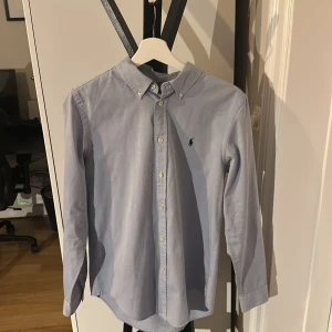 Ljusblå skjorta från Ralph Lauren - Klassisk ljusblå skjorta från Ralph Lauren med button-down krage och broderad logga på bröstet. Skjortan har långa ärmar och knäppning framtill. Perfekt för en stilren och tidlös look.