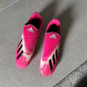 Adidas rosa fotbollsskor med svarta detaljer - Säljer ett par Adidas fotbollsskor i en riktigt snygg rosa färg med svarta ränder och vita detaljer. Skorna har en strumpliknande passform utan snörning och fasta dobbar för spel på gräs. Perfekta för dig som vill sticka ut på planen! Skorna är använda men väldigt bra skick och nytvättade! Storlek45,1/3
