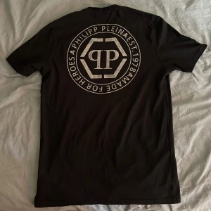 Philipp Plein - Säljer en svart t-shirt från Philipp Plein med en stor dödskalle framtill i rhinestone mönster och logga både fram och bak. T-shirten har rund hals och korta armar.