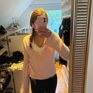 Ljusrosa omlottkofta med knyt - Supermjuk ljusrosa omlottkofta från Gina Tricot med knyt i sidan och v-ringning. Tröjan har långa ärmar och är stickad i ett fluffigt material som känns riktigt mysigt. Koftan är knappt använd 