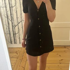 Svart kortklänning med knappar - Svart kortklänning från H&M med v-ringning och korta puffärmar. Klänningen har dekorativa knappar framtill och en figurnära passform som ger en snygg siluett. Perfekt för dig som gillar enkel men stilren design.