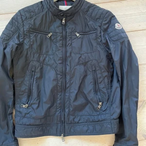 Svart vindjacka från Moncler - Tja, säljer dena snygga svarta vindjacka från Moncler med fyra dragkedjefickor framtill och quiltade detaljer på axlarna. Jackan har hög krage och långa ärmar, perfekt för blåsiga dagar. Klassisk och stilren design med Moncler-logga på ärmen. 100% riktig
