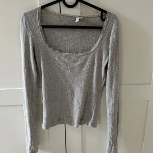 Grå långärmad topp med spets från Nelly  - Säljer en grå långärmad topp från Nelly med rundad, djup urringning och fina spetsdetaljer längs kanten. Tunt, ribbat material som är mjukt och stretchigt. Perfekt att styla till jeans eller kjol.
