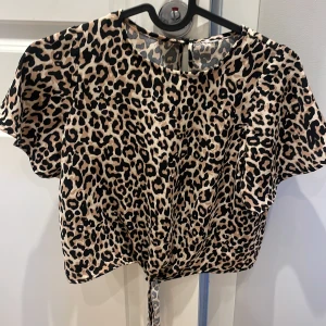 Leopardmönstrad blus med knyt - Säljer en snygg blus med leopardmönster och korta ärmar. Blusen har rund halsringning och knyt i midjan för en cool detalj. Perfekt för dig som gillar djurmönster och vill sticka ut.