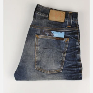 Vintage Blå jeans från Nudie Jeans - Snygga blå jeans från Nudie Jeans med klassisk femficksdesign och orangea kontrastsömmar. Jeansen har knäppning med knappar och en rak passform. Slitningar och tvättade detaljer ger en cool, använd look. Blev tyvärr försmå för min smak. Stolek 31:32
