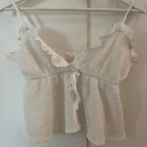 Vit croppad linnetopp med volangdetaljer - Säljer en söt vit croppad linnetopp med smala axelband och volangdetaljer längs kanterna. Toppen har knytning framtill och smockad struktur som ger en bekväm passform❤️