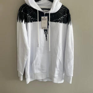 Marcelo Burlon tröja - Säljer en vit hoodie från Marcelo Burlon med ett svart vingmönster över axlar och bröst. Tröjan har huva med dragsko och en stor magficka framtill. Perfekt statement-plagg för dig som gillar unika detaljer.