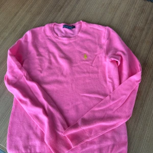 Rosa tröja från Ralph Lauren - Säljer en snygg rosa tröja från Ralph Lauren med det klassiska logotypbroderiet i gult på bröstet. Tröjan har långa ärmar och en rund hals.