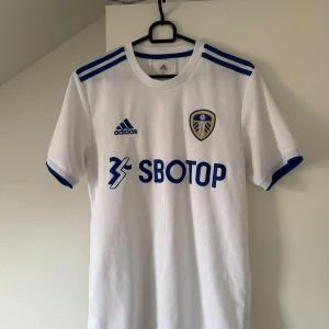 Vit fotbollströja från Leeds - Säljer en vit fotbollströja från Adidas med blå detaljer och korta ärmar. Tröjan har klubbmärke och sponsorlogga på framsidan. Perfekt för fotbollsfans som vill visa sitt stöd. Tillverkad i AEROREADY-material för optimal komfort. Passar även M
