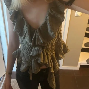 Grön volangblus - Säljer en snygg grön blus med volanger och djup v-ringning. Perfekt för att ge en romantisk touch till din outfit. Blusen har en knytning framtill och är i ett lätt och luftigt material.