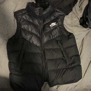 Svart dunväst från Nike - Säljer en svart dunväst från Nike med dragkedja och hög krage. Västen har en klassisk design med Nike-logga på bröstet. Perfekt för kyligare dagar.
