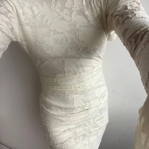 Vit spetsklänning från Boohoo - Säljer en elegant vit spetsklänning från Boohoo med öppen rygg och långa ärmar. Klänningen har en tight passform och är perfekt för speciella tillfällen. Den har ett vackert blommigt mönster och en kort längd.