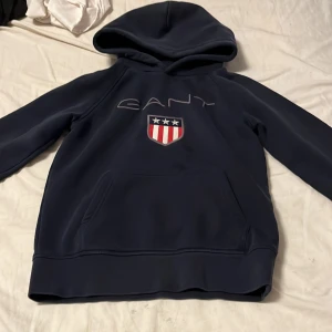 Mörkblå hoodie från GANT - Säljer en snygg mörkblå hoodie från GANT med broderad logga och emblem på bröstet. Perfekt för en avslappnad stil. Den har en klassisk känguruficka och en bekväm huva.
