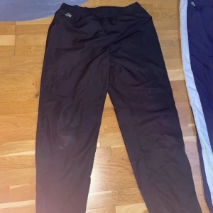 Svarta träningsbyxor från Lacoste - Säljer ett par svarta träningsbyxor från Lacoste Sport. Byxorna har en elastisk midja med snörning och är perfekta för sportiga aktiviteter. Den klassiska Lacoste-loggan syns på sidan.