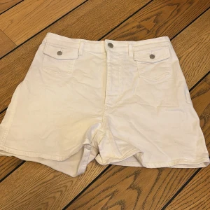 Vita shorts med fickor - Säljer ett par vita shorts med hög midja och praktiska fickor fram och bak. De har en knapp och dragkedja framtill för enkel stängning. Perfekta för varma sommardagar!