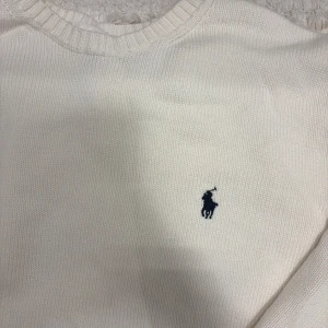 Vit stickad tröja från Ralph Lauren - Säljer en klassisk vit stickad tröja från Ralph Lauren med deras ikoniska logga broderad på bröstet. Tröjan har långa ärmar och ribbstickade muddar vid ärmslut och nederkant. Perfekt för en stilren look.