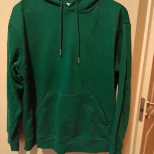 Grön hoodie - Säljer en snygg grön hoodie med känguruficka och justerbar huva. Perfekt för en avslappnad stil och bekväm att bära under kyligare dagar. Passar bra till jeans eller mjukisbyxor.