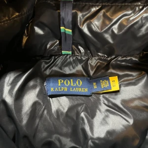 Svart dunjacka från Ralph Lauren - Säljer en snygg svart dunjacka från Ralph Lauren. Jackan har en glansig finish och är utrustad med en huva och dragkedja! Pris kan diskuteras 