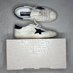 Vita sneakers med svart stjärna - Säljer ett par vita sneakers från Golden Goose med en svart stjärna på sidan. Skorna har snörning och en något sliten look som ger dem en unik stil. Perfekta för en avslappnad och trendig look.