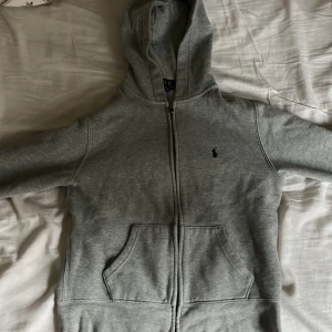 Grå hoodie från Ralph Lauren - Säljer en stilren grå hoodie från Ralph Lauren med dragkedja och klassisk logga på bröstet. Tröjan har en bekväm passform med långa ärmar och praktiska fickor framtill. Perfekt för en avslappnad look.