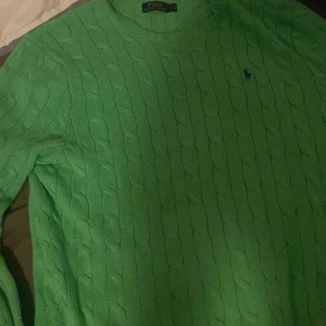 Grön stickad tröja från Ralph Lauren - Säljer en snygg grön stickad tröja st L från Ralph Lauren med kabelstickat mönster. Tröjan har långa ärmar och en klassisk rund halsringning. Perfekt för en stilren look. 