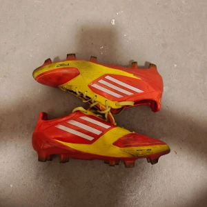 Röda och gula fotbollsskor från Adidas - Säljer ett par röda och gula fotbollsskor från Adidas med vita ränder och snörning. Perfekta för fotbollsplanen med sin sportiga design och slitstarka material.