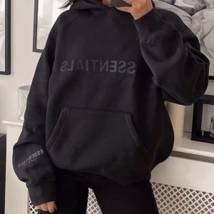 Svart hoodie från Essentials - Säljer en svart hoodie från Essentials med tryck på bröstet och ärmen. Den har en klassisk känguruficka och en bekväm passform. Perfekt för en avslappnad stil.