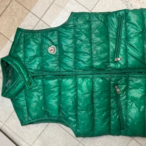 Grön dunväst från Moncler - Säljer en snygg grön dunväst från Moncler med dragkedja och två praktiska fickor framtill. Skick:9/10 Liten repa på kragen men inget man tänker på Passar M/S