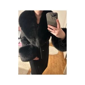 Svart kashmir kappa med äkta päls  - Elegant svart kappa med lyxiga pälsdetaljer på krage och ärmslut. Perfekt för att hålla sig varm och stilfull under kyliga dagar. Kappan har en klassisk design med en figursydd passform och knäppning framtill.