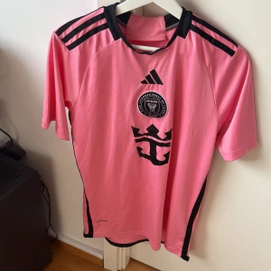 Rosa fotbollströja från Adidas - Snygg rosa fotbollströja från Adidas med svarta detaljer och korta ärmar. Tröjan har ett tryck med ett emblem på framsidan och texten 'Messi 10' på baksidan. Perfekt för fotbollsfans som vill sticka ut på planen eller läktaren.