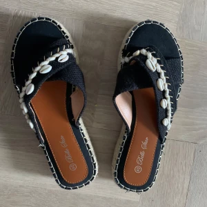 Svarta sandaler med snäckdetaljer från Bella Star - Snygga svarta sandaler från Bella Star med dekorativa snäckor och flätad sula. Perfekta för sommarens alla äventyr. De har en bekväm passform och stilren design. Strl 40 men små så är som 39