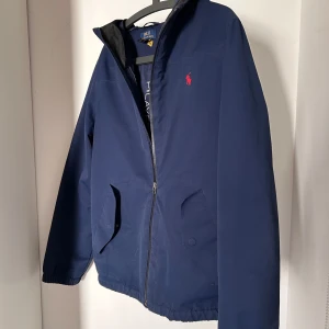 Blå jacka från Polo Ralph Lauren - Snygg blå jacka från Polo Ralph Lauren med dragkedja och huva. Jackan har en röd broderad logotyp på bröstet och praktiska fickor med knappar. Perfekt för en stilren look. Utmärkt jacka under vår/sommar tiderna som väntas komma. Köpt inför sommaren 2024 men använd sparsamt och därför fortfarande som ny utan någon synlig skada av något slag. XL Barnstorlek, passar 165cm - ca 177cm. Jackan är vind och regntålig.                                                                  Hör av er vid frågor!
