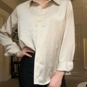 Beige sidenblus - Elegant beige blus i siden med klassisk krage och knappar framtill. Blusen har långa ärmar med knappar vid ärmsluten, vilket ger en stilren look. Perfekt för en sofistikerad stil.