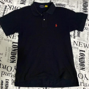 Svart pikétröja från Ralph Lauren - Säljer en klassisk svart pikétröja från Ralph Lauren med den ikoniska röda loggan på bröstet. Tröjan har korta ärmar och en traditionell krage med knappar. Perfekt för en stilren look.