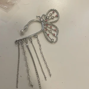 Silverfärgad ördekoration med pärlor och kedjor - Elegant ördekoration i silverfärg formad som en fjäril med glittriga detaljer och pärlor. Den har flera hängande kedjor och pärlor som ger en unik look. Perfekt för att ge en extra touch till din frisyr.