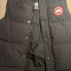 Svart dunväst från Canada Goose - Säljer en svart dunväst från Canada Goose med deras ikoniska logga på bröstet. Västen har en dragkedja och knappar framtill för extra skydd mot kyla. Perfekt för kyliga dagar när du vill hålla värmen utan att kompromissa med stilen. Kvitto finns.