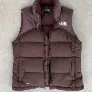 Brun dunväst från The North Face - Säljer en brun dunväst från The North Face med dragkedja och hög krage. Västen har märkets logga på bröstet och ryggen. Perfekt för kyligare dagar.