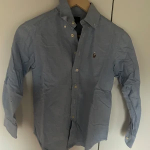 Blå skjorta från Ralph Lauren - Snygg blå skjorta från Ralph Lauren med klassisk krage och knappar framtill. Skjortan har långa ärmar och en liten broderad logotyp på bröstet. Perfekt för en stilren look.