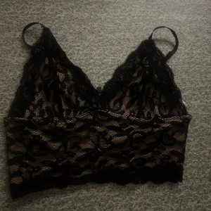 Svart spets bralette - Snygg svart bralette med spetsdetaljer och justerbara axelband. Den har en beige underton som ger en fin kontrast till spetsen. Perfekt för att lägga till en touch av elegans till din outfit.