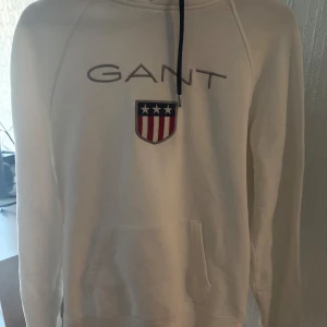 Vit hoodie från GANT - Säljer en stilren vit hoodie från GANT med ett tryck av märkesnamnet och en sköld med stjärnor och ränder på bröstet. Hoodien har en klassisk passform med långa ärmar och en praktisk magficka. Perfekt för en avslappnad look.