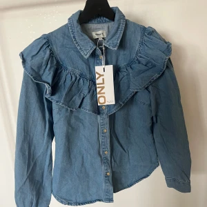 Blå jeansskjorta från ONLY - Snygg blå jeansskjorta från ONLY med volangdetaljer över axlarna och knappar framtill. Aldrig använd då jag skulle returnera den p.g.a för liten storlek men glömde av att returnera i tid 