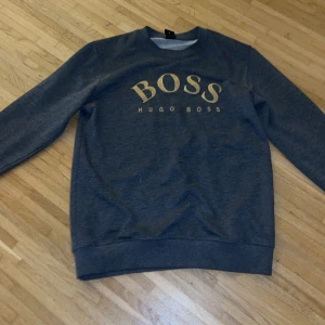 Grå sweatshirt från Hugo Boss - Säljer en stilren grå sweatshirt från Hugo Boss med BOSS-logga i guld på bröstet. Tröjan har långa ärmar och en klassisk rund halsringning. Perfekt för en avslappnad och trendig look.