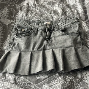 Grå jeanskjol med volang - Snygg grå jeanskjol med volangdetalj längst ner. Kjolen har klassiska femficksdetaljer och en knapp- och dragkedjestängning framtill. Perfekt för en trendig look!