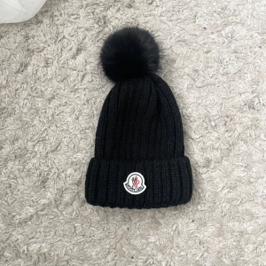 Svart stickad mössa med pälsboll - Snygg svart stickad mössa från Moncler med en stor pälsboll på toppen. Mössan har en uppvikt kant och ett broderat märke framtill. Perfekt för kyliga dagar!