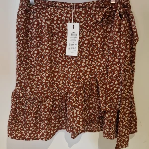 Brun blommig kjol med volang - Snygg brun kjol från ONLY blommigt mönster och volangdetaljer. Kjolen har en omlott-design med knytning i midjan och är tillverkad i 100% polyester. Perfekt för en trendig look!