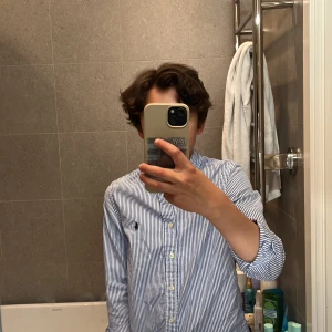 Hej jag seljer en polo Ralph Lauren - Snygg blå- och vitrandig skjorta med en liten broderad logga på bröstet. Skjortan har långa ärmar och knappar framtill. Perfekt för en stilren look.