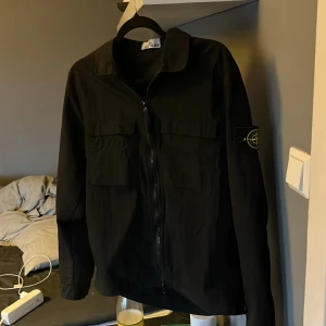 Svart jacka från Stone Island windbreaker - Säljer en stilren svart jacka från Stone Island. Jackan har en dragkedja framtill och två bröstfickor. Den är långärmad och har en klassisk krage. Perfekt för en cool och avslappnad stil.