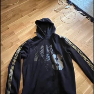 Svart hoodie från Plein Sport - Snygg svart hoodie från Plein Sport med ett coolt tigertryck och nummerdetalj på framsidan. Långärmad med märkets logga längs ärmarna. Perfekt för en sportig look!