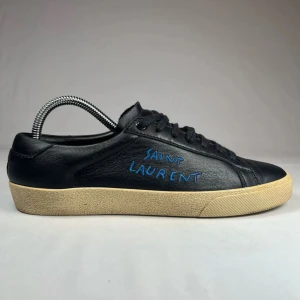 Svarta sneakers från Saint Laurent - Snygga svarta sneakers från Saint Laurent med blå logotyp på sidan. Skorna har en rund tå och snörning framtill. De har en beige sula som ger en cool kontrast till det svarta skinnet.