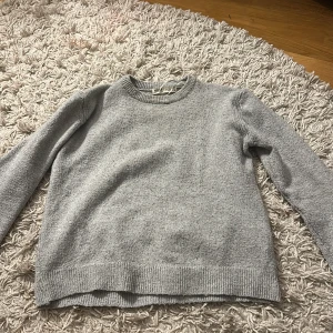 Grå stickad tröja - Säljer en mysig grå stickad tröja med rund hals och långa ärmar. Perfekt för kyligare dagar. Tröjan har ribbstickade muddar vid ärmslut och nederkant.